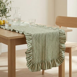 May include: A green and white striped table runner with ruffled edges. The runner is placed on a light wood table, with glasses and a water pitcher visible. The runner adds a touch of elegance to the dining table.