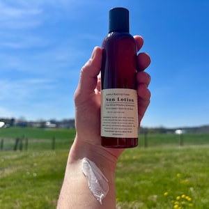 May include: A brown bottle of Simply Rooted Farm Sun Lotion held against a blue sky. The label lists ingredients and product details. A dollop of white lotion is on the arm. The background shows a green field and trees.