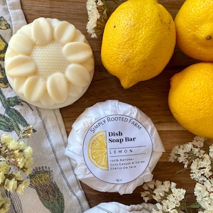May include: A white dish soap bar wrapped in white paper with a yellow lemon slice and the text "Simply Rooted Farm Dish Soap Bar Lemon 100% Natural Cuts Grease & Grime Gentle on Skin 4 oz" on the front. The soap bar is surrounded by lemons and white flowers.
