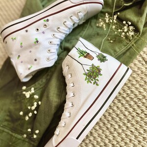 Puede incluir: Zapatillas blancas de lona de caña alta con ribete rojo. Un zapato tiene un diseño floral, el otro tiene un diseño pintado a mano de plantas en macetas colgando de una cuerda.