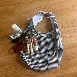May include: A black and white gingham hobo bag with a white ribbon tie at the top and a front pocket. The bag contains a tube of shea butter hand cream, a lipstick, a small bottle, a hair scrunchie, sunglasses, and a hair clip.