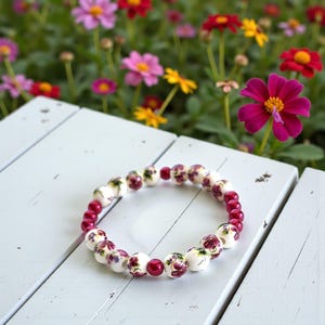 May include: A beaded bracelet featuring white ceramic beads with floral designs and deep red beads. The bracelet is resting on a white wooden surface, with colorful flowers in the background. The bracelet is approximately 7 inches in circumference.
