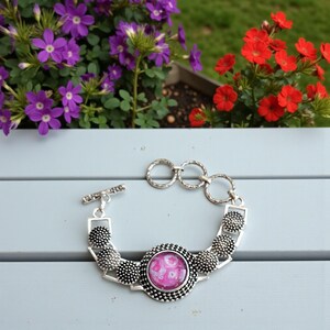 May include: Silver-toned bracelet with a central pink and white patterned stone. The bracelet features a toggle clasp and decorative elements with a dotted texture. The background includes purple and red flowers.
