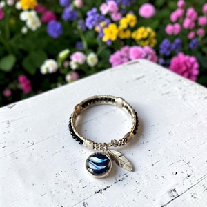 May include: A circular bracelet featuring a band of small black beads and metallic accents. The bracelet includes two dangling charms: a round pendant with a dark blue and white swirled pattern and a silver-toned metal feather charm.