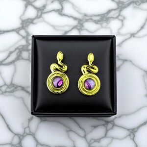 Peut inclure: Une paire de boucles d'oreilles serpent dorées avec des centres violets, présentées sur une boîte carrée noire. Les boucles d'oreilles présentent un motif de serpent enroulé au-dessus d'une pierre violette circulaire, sertie dans un cadre doré texturé.