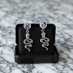 May include: A pair of silver-colored snake earrings with blue and white marbled accents. The earrings are displayed in an open black jewelry box on a marble surface. The snake design is detailed and coiled.