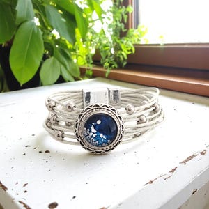 May include: White leather-like wrap bracelet with a silver magnetic clasp and decorative silver beads. The focal point is a round, silver-toned charm with a dark blue and white design, surrounded by a decorative silver border. A stylish accessory.