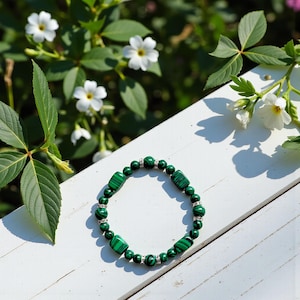 May include: A green malachite bracelet with a mix of round and rectangular beads, interspersed with small, silver-colored spacers. The bracelet is displayed on a white surface, with white flowers and green foliage in the background.