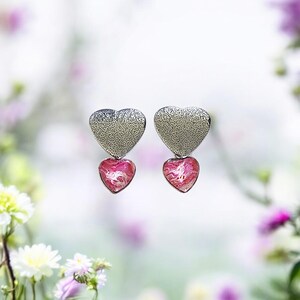 May include: A pair of silver heart-shaped earrings with pink heart-shaped accents. The earrings have a textured surface.