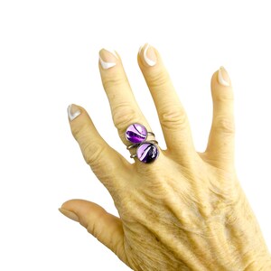 May include: Two silver rings with round, purple and black abstract designs. The rings are worn on a hand with manicured nails. The rings have a modern, artistic style and are set against a white background.