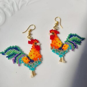 May include: A pair of beaded rooster earrings with a rainbow coloured tail and a red, orange, and yellow body. The earrings are made with small beads and have gold hooks.