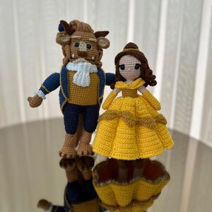 May include: Handmade crochet figures of the Beast and Belle from Beauty and the Beast. The Beast is in a blue and gold suit with a white cravat. Belle wears a yellow dress. The figures are crafted with a whimsical style.