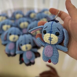 May include: Crocheted blue donkey keychain with a light yellow muzzle, pink inner ears, and a purple belly. The keychain is held in a hand, with several similar keychains in the background.