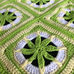 Pode incluir: Close-up de um cobertor de crochê com um padrão repetitivo de flores verdes e brancas. O cobertor é feito com um fio macio e texturizado.