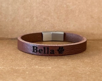 Puede incluir: Un brazalete de cuero marrón con cierre magnético. El brazalete está grabado con el nombre "Bella" y una huella de pata.