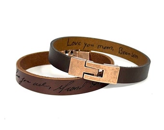 May include: Two brown leather bracelets with a copper clasp. One bracelet is engraved with the text "Love you mom, Branson". The other bracelet is engraved with the text "Love you always" and a handwritten signature.