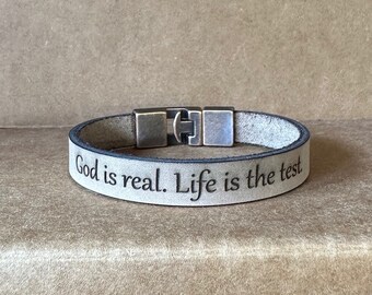Puede incluir: Un brazalete de cuero marrón claro con un cierre de plata. El brazalete tiene el texto "God is real. Life is the test." grabado.