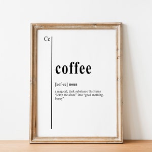 Peut inclure: Impression encadrée avec la définition du mot "coffee" en noir sur fond blanc. La définition décrit le café comme une substance magique. Le cadre est de couleur marron clair, et l'impression est posée sur une surface en bois.