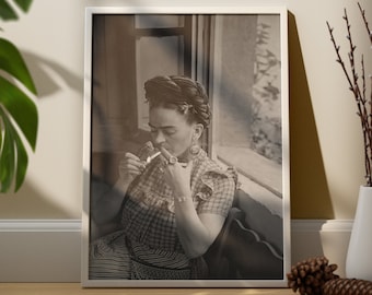 Può includere: Una stampa ritratto in bianco e nero incorniciata che raffigura Frida Kahlo con i capelli raccolti in uno chignon, che indossa una camicetta a motivi e gioielli, mentre tiene in mano una sigaretta accesa. L'opera è in una cornice bianca.