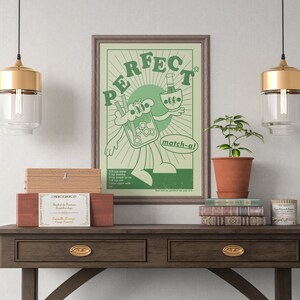 May include: A framed green and white poster with the word "PERFECT" and a cartoon character holding a matcha drink. The poster is on a dark wood table with drawers, along with a plant, books, and decorative boxes.