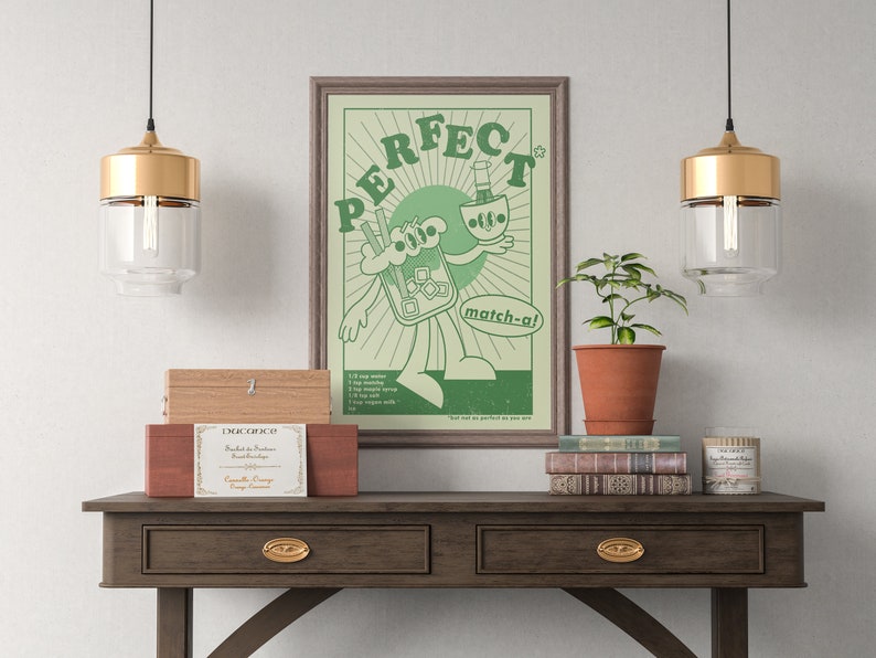May include: A framed green and white poster with the word "PERFECT" and a cartoon character holding a matcha drink. The poster is on a dark wood table with drawers, along with a plant, books, and decorative boxes.