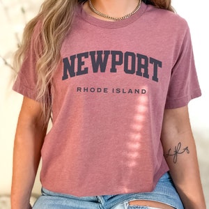 以下が含まれることがあります： マウブ色のクロップドTシャツに、黒い文字で「NEWPORT RHODE ISLAND」と書いてあります。