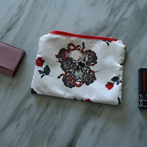 May include: A white zippered pouch with a skull and snake design surrounded by red roses. A pink wallet and a black lipstick tube are also pictured.