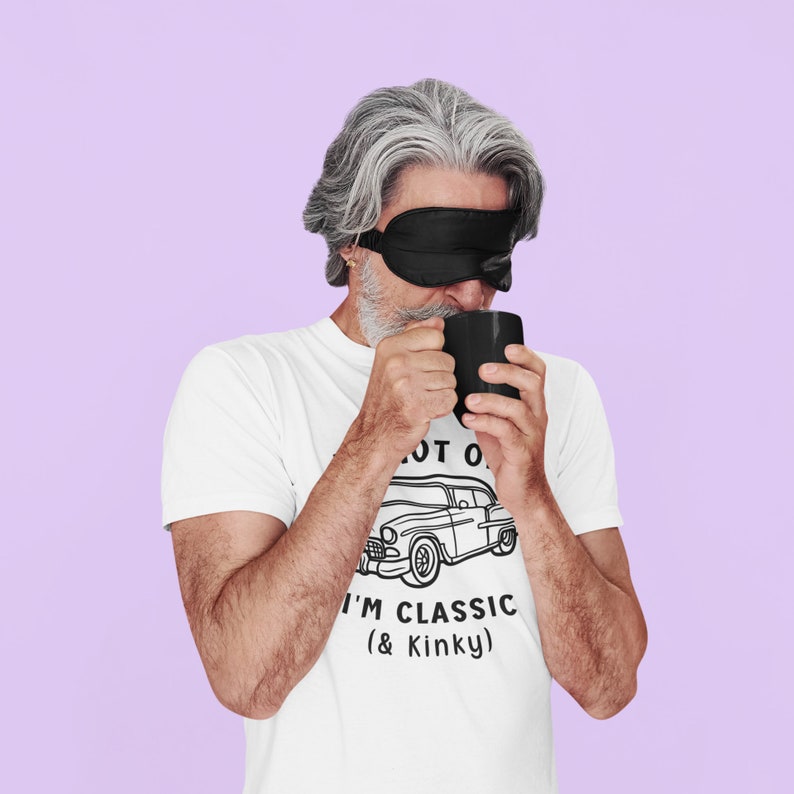 K&ouml;nnte beinhalten: Ein Mann mit einer schwarzen Schlafmaske trinkt aus einer schwarzen Tasse. Er tr&auml;gt ein wei&szlig;es T-Shirt mit einer schwarz-wei&szlig;en Illustration eines Autos und dem Text "I'm Classic (& Kinky)".