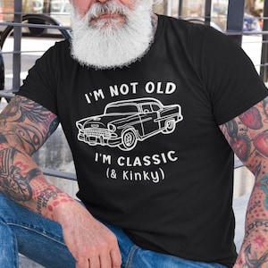 K&ouml;nnte beinhalten: Ein schwarzes T-Shirt mit wei&szlig;em Text, der "I'm not old I'm classic (& Kinky)" lautet. Oberhalb des Textes befindet sich eine Illustration eines Oldtimers. Die Person, die das Shirt tr&auml;gt, hat T&auml;towierungen auf den Armen.