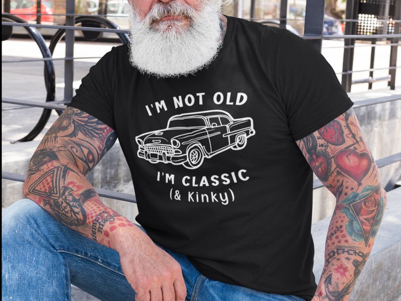 K&ouml;nnte beinhalten: Ein schwarzes T-Shirt mit wei&szlig;em Text, der "I'm not old I'm classic (& Kinky)" lautet. Oberhalb des Textes befindet sich eine Illustration eines Oldtimers. Die Person, die das Shirt tr&auml;gt, hat T&auml;towierungen auf den Armen.
