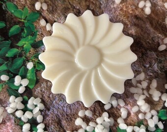 May include: A white, round, decorative soap bar with a flower-like design. The soap is surrounded by white wax beads.