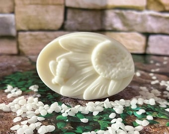 May include: A white oval-shaped soap bar with a raised design of a flower and a bee. The soap is sitting on a surface covered in white rice-shaped granules.