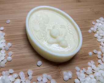 May include: A white oval-shaped soap bar with a raised design of a bee and a flower. The soap is on a wooden surface with white wax pellets scattered around it.