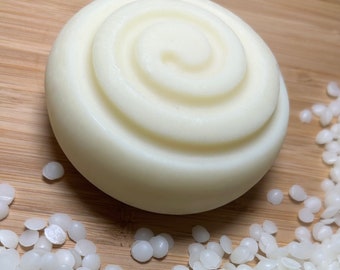 May include: A white, round, solid bar of soap with a spiral design on the top. The soap is sitting on a wooden surface with white beads scattered around it.