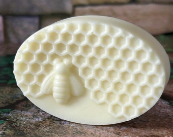 May include: Oval-shaped, off-white soap bar with a detailed honeycomb pattern and a bee relief. The soap has a smooth surface and a natural, handcrafted appearance, perfect for a spa-like experience.