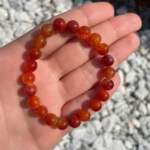 Puede incluir: Un brazalete de cuentas de ágata roja con un acabado de piedra natural. Las cuentas son redondas y tienen una apariencia ligeramente translúcida.
