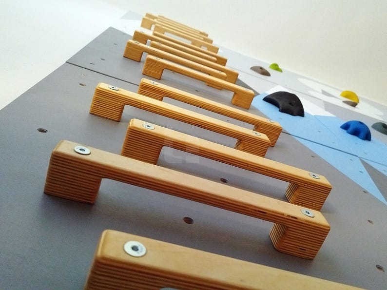 K&ouml;nnte beinhalten: Nahaufnahme einer Kletterwand mit Holzgriffen. Die Griffe sind versetzt angeordnet und aus hellem Holz gefertigt. Die Wand ist hellgrau gestrichen.