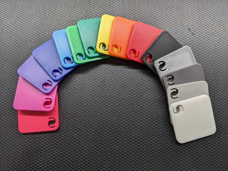 May include: A rainbow arc of colorful, square-shaped plastic tags. The tags are in shades of red, pink, purple, blue, green, yellow, orange, black, gray, and white. Each tag has a textured surface and a unique cutout design.