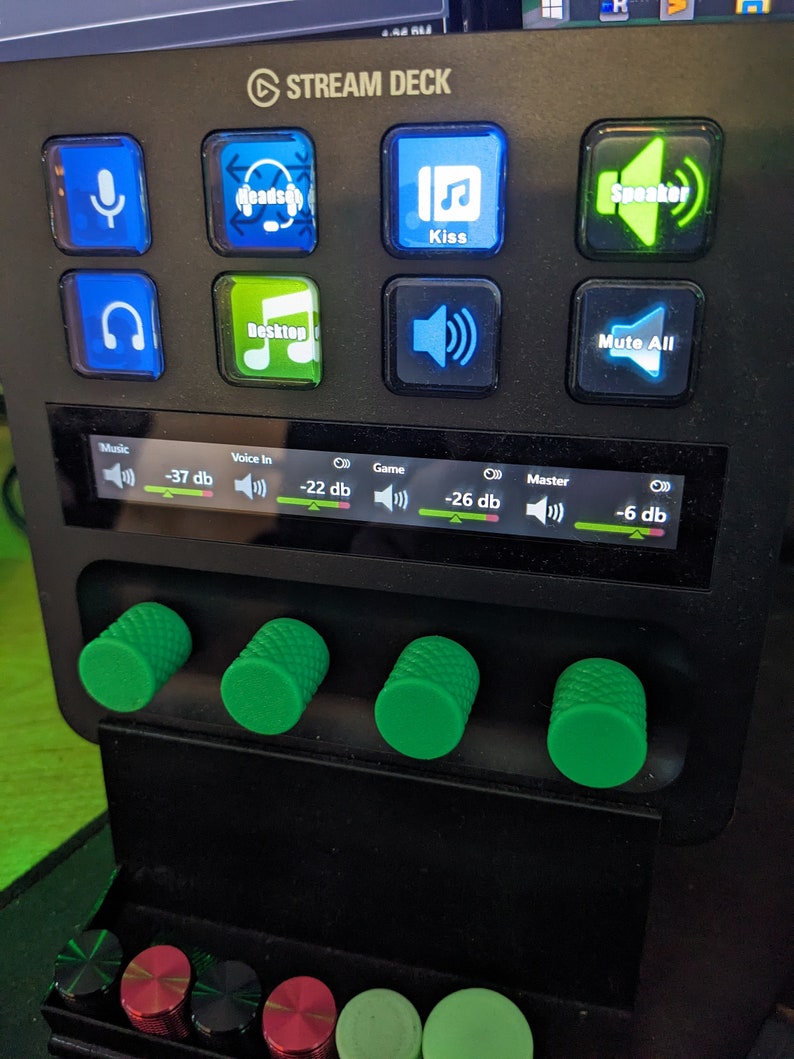 May include: A black Stream Deck control panel with illuminated buttons. The buttons display icons for a microphone, headphones, music notes, and speaker. Green knobs and colored dials are visible at the bottom. The screen displays audio levels.