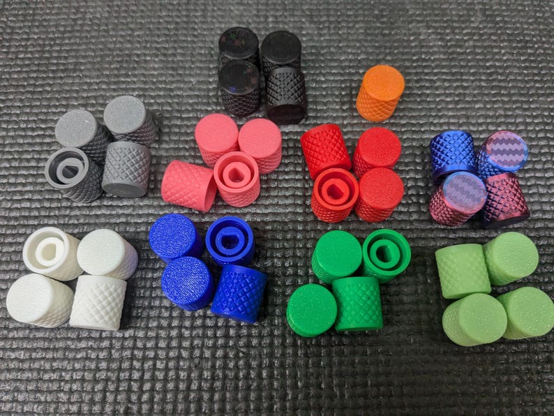 May include: A collection of colorful, cylindrical knobs in various colors, including black, gray, pink, red, orange, blue, green, and white. Some knobs have a textured diamond pattern, while others are smooth. The knobs are arranged on a textured gray surface.