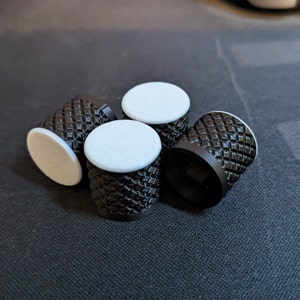 May include: Four black knobs with a diamond pattern on the sides and white circular tops are arranged on a dark surface. The knobs appear to be made of metal or a similar material.