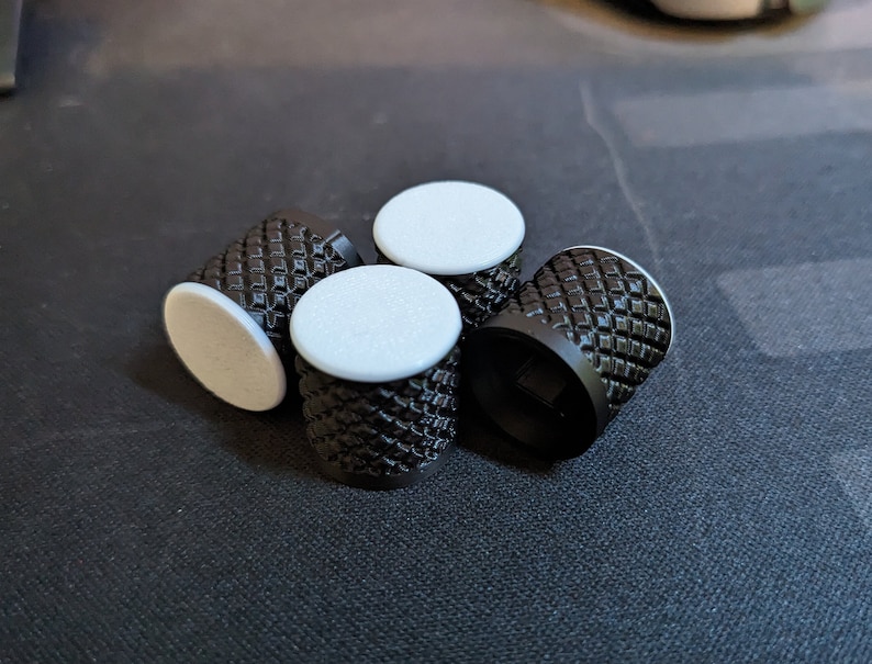 May include: Four black knobs with a diamond pattern on the sides and white circular tops are arranged on a dark surface. The knobs appear to be made of metal or a similar material.