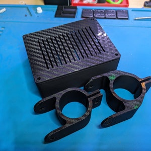 以下が含まれることがあります： カーボンファイバー模様の黒い3Dプリントクランプが2つ。クランプは、カーボンファイバー模様のトップを持つ長方形のボックスを保持するように設計されています。