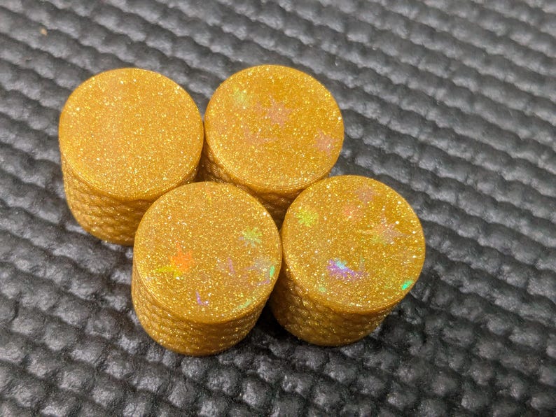 May include: Four stacks of golden, glittery, round objects. Each stack is composed of several circular pieces. The objects have a textured appearance and display iridescent flecks of color, creating a shimmering effect. The background is a dark, textured surface.