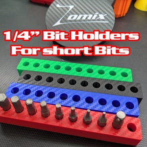 Puede incluir: Una pila de soportes para puntas de 1/4" en rojo, azul, negro y verde, con el texto "1/4" Bit Holders For short Bits". Los soportes están diseñados para organizar puntas cortas. Fabricados en fibra de carbono.