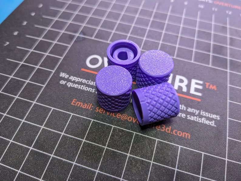 May include: Four purple, textured knobs with a diamond pattern on the sides and a flat, textured top. One knob is disassembled, revealing the interior. The knobs are on a black surface with a grid pattern and the word "OVERTURE" is visible.