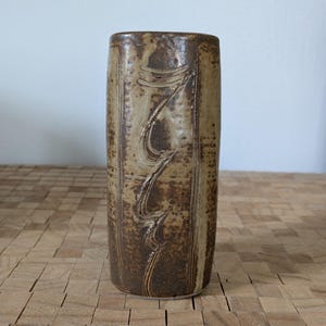 May include: A tall, cylindrical ceramic vase with a textured, earthy brown and tan finish. The vase features vertical lines and a central, stylized wave design. The handmade pottery is suitable for home decor.