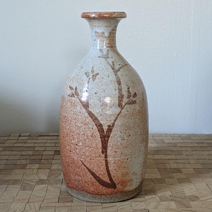 May include: A ceramic vase with a speckled, light brown and white finish. The vase features a slender neck and a rounded body. A brown, stylized floral design decorates the front. The vase is approximately 10 inches tall.