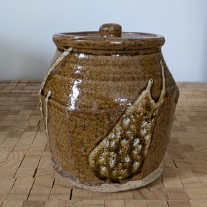 May include: A brown ceramic jar with a lid, featuring a white glaze and a speckled design. The jar has a rounded body and a slightly flared opening.