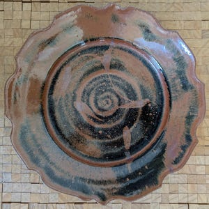 May include: A large, round ceramic plate with a scalloped edge. The plate has a brown and black glaze with a swirling pattern.